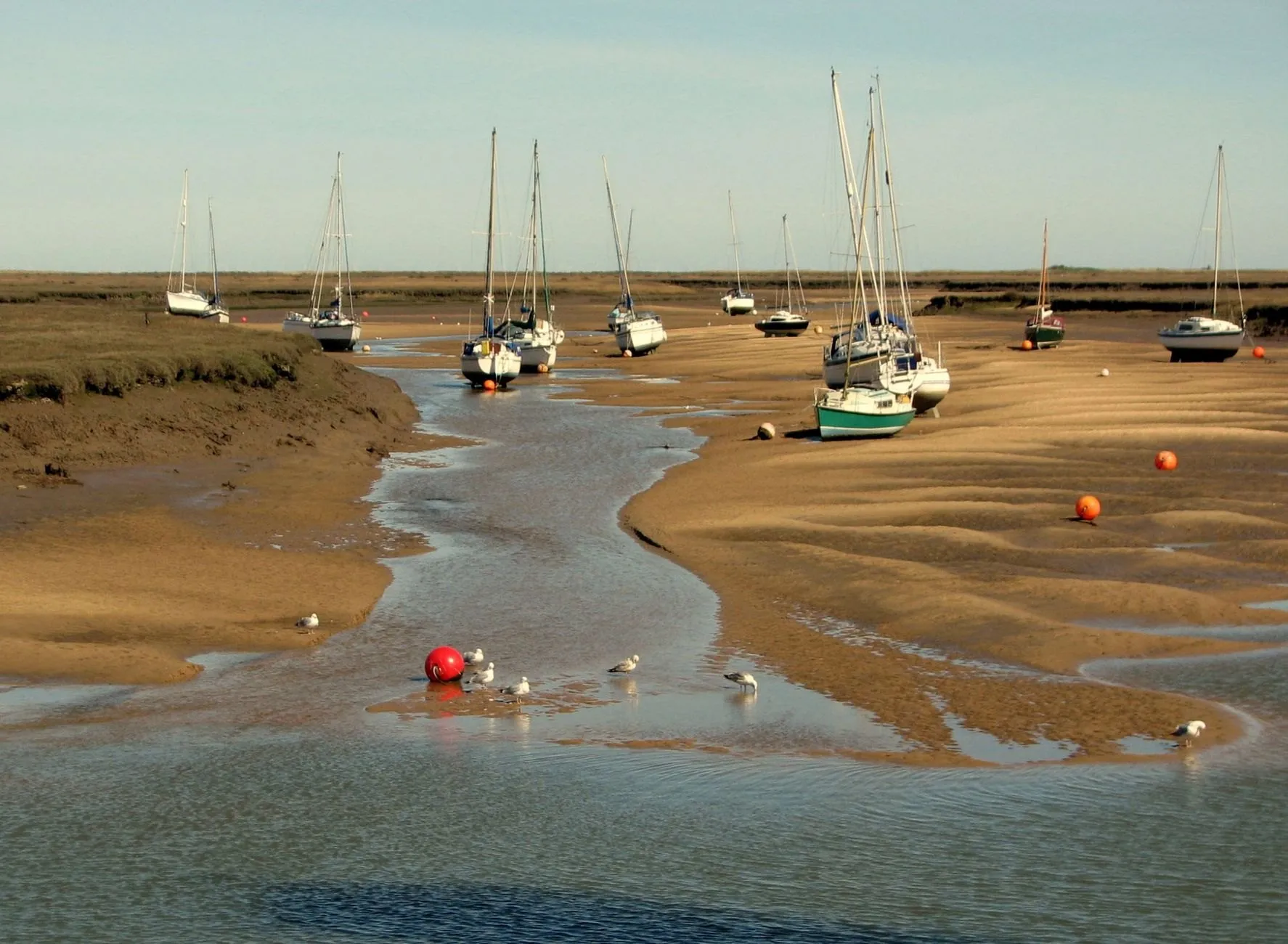 Yachts resting on sandbanks either side of a shallow channel at low tide, with red mooring buoys