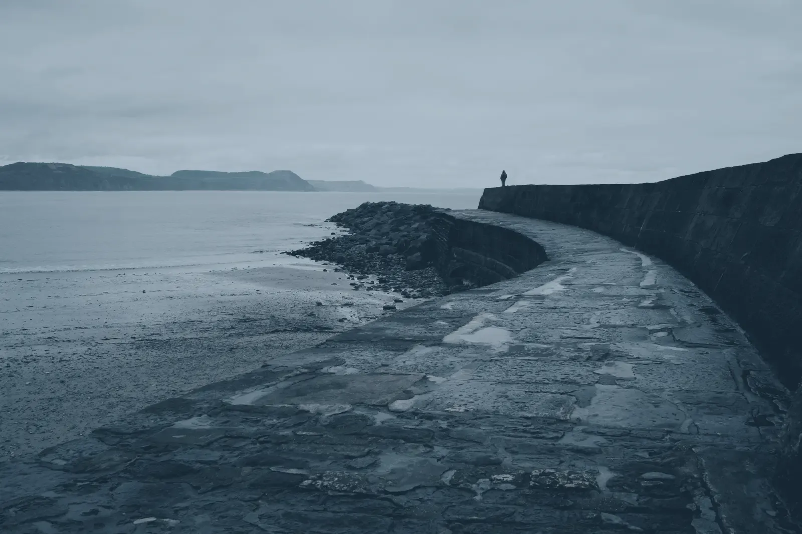 A long curving stone harbour wall reaching out over ground that dries, one figure at the far end.